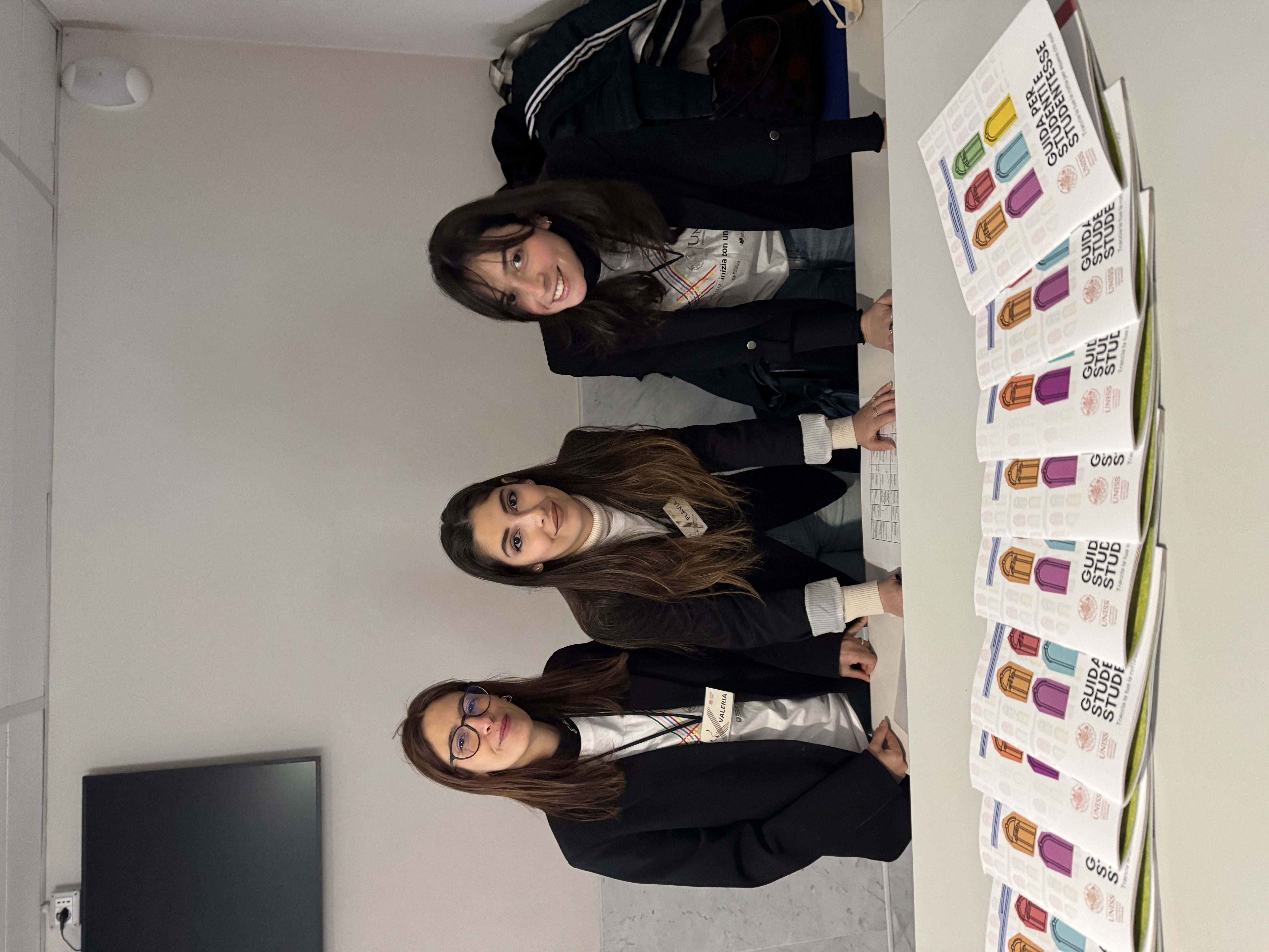 Tre componenti dello staff UNISS posano dietro il desk informativo con guide per studenti durante l’accoglienza all’Università di Sassari